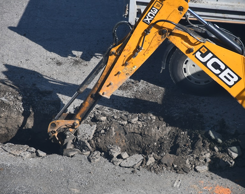 Yellow JCB excavator arm breaking asphalt road on a construction site.