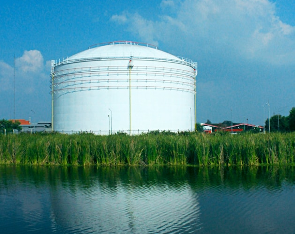 Large white industrial storage tank behind green grass, reflected in nearby water under a blue sky.